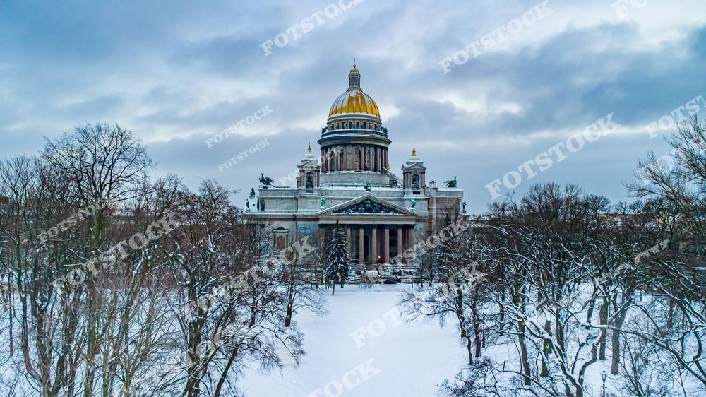 Фотография зимнего Санкт-Петербург, Россия. На фото изображена панорама зимнего Петербурга с Исаакиевский собор, окружённым заснеженными деревьями. Золотой купол собора эффектно выделяется на фоне серого зимнего неба, создавая выразительный контраст. Фотоснимок передаёт атмосферу зимы в России и спокойное величие Исаакиевская площадь. Это изображение подчёркивает красоту архитектуры Санкт-Петербурга в холодное время года и может использоваться как иллюстрация, картинка или слайд, посвящённые зимним городским пейзажам.